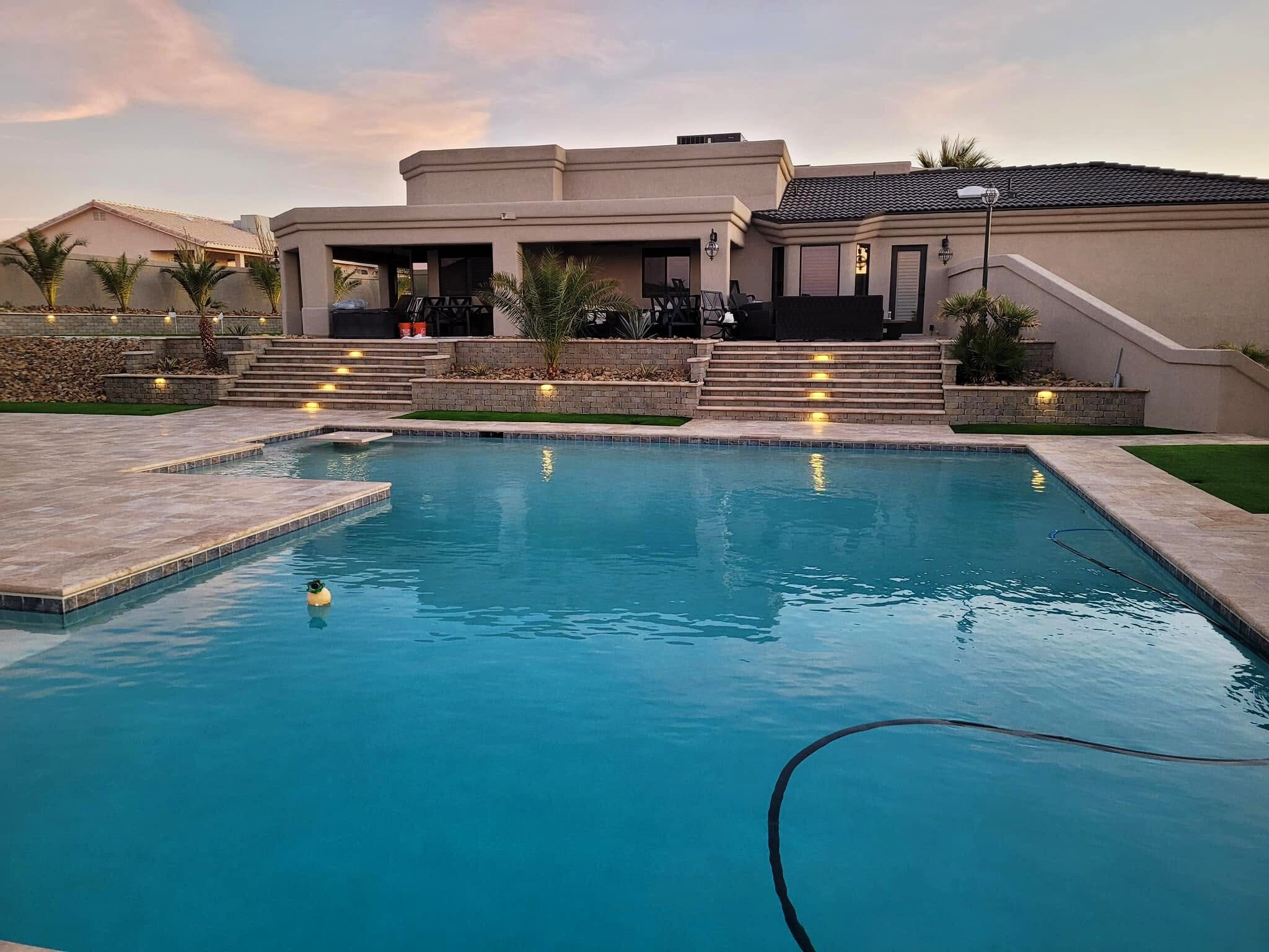 Finished travertine pool deck with tiered lit retaining walls and palms at dusk — a Havasu Pavers backyard in Lake Havasu City
